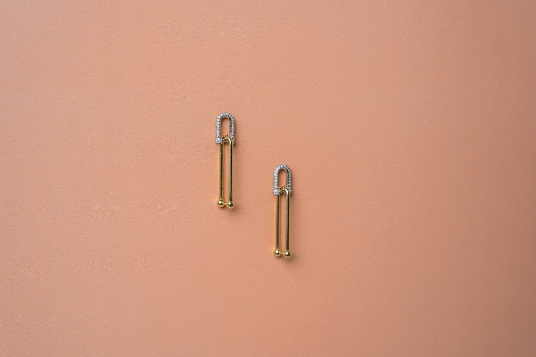 To guldfarvede, aflange smykker med sølvfarvede detaljer på rosa baggrund